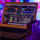 A macBook displaying an audio mixing program sits on a table next to other audio equipment in a night club atmosphere